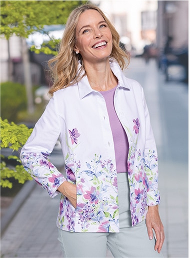 Woman smiling over the camera, wearing a floral print jackets over a purple shirts and light pants, with one hand in the jacket pocket