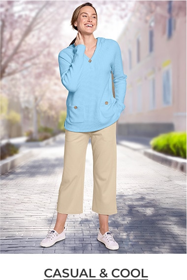 woman smiling, standing outside a house with one hand in her pocket, wearing blue cardigans and tan pants