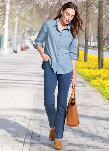 woman walking down a paved path between trees, ooking down and wearing jeans shirt and blue jean pants, carrying a brown bag