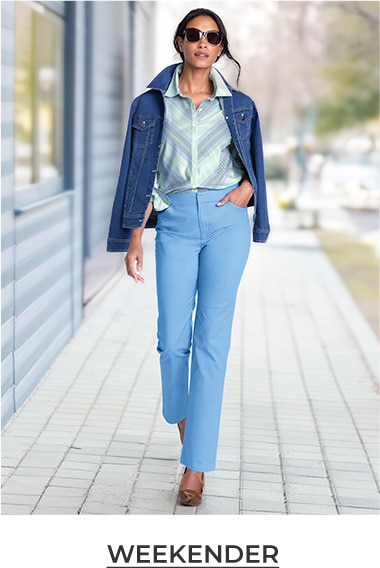 woman walking toward the camera wearing a light green, striped shirt and blue jeans, with a jeans jackets over her shoulders and sunglasses on