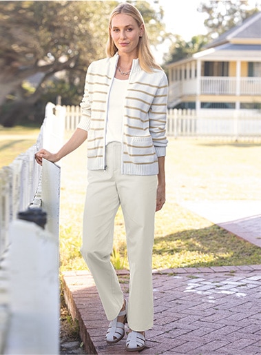 woman smiling into the camera, standing outside a house with one hand on a white fence, wearing light colored cardigans, shirt, and pants