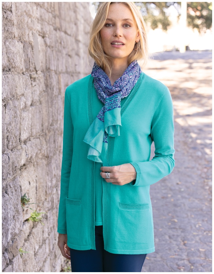 women standing on a paved path and facing the camera, wearing a green cardigan, purplr scarf, and blue jeans