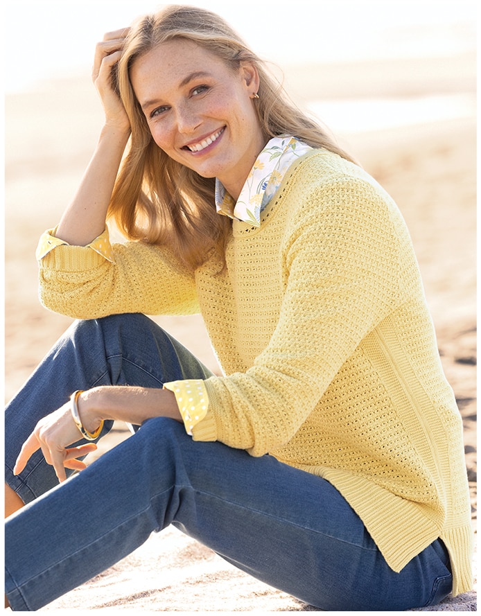 woman sitting on a beach wearing a yellow sweater and blue jeans