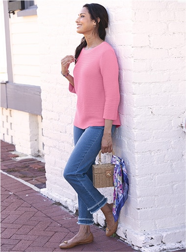 woman facing to the left, leaning against a wall wearing a pink sweaters and blue jeans