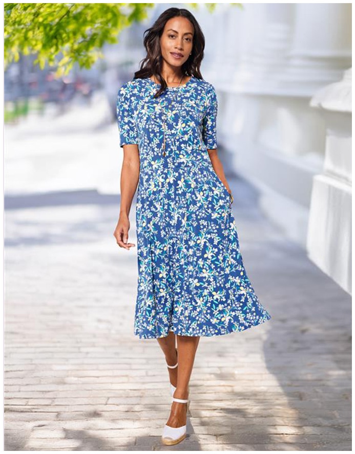 woman smiling into camera, wearing a light blue floral print dress and white shoes