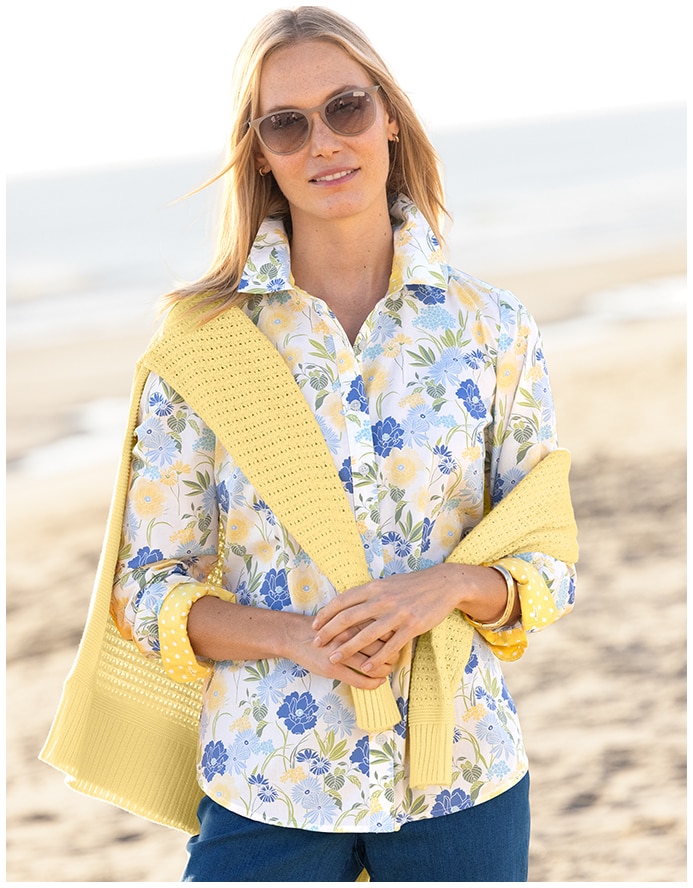 women on a beach, facing the camera, wearing a floral shirt with yellow sweater draped over her shoulders and blue jeans